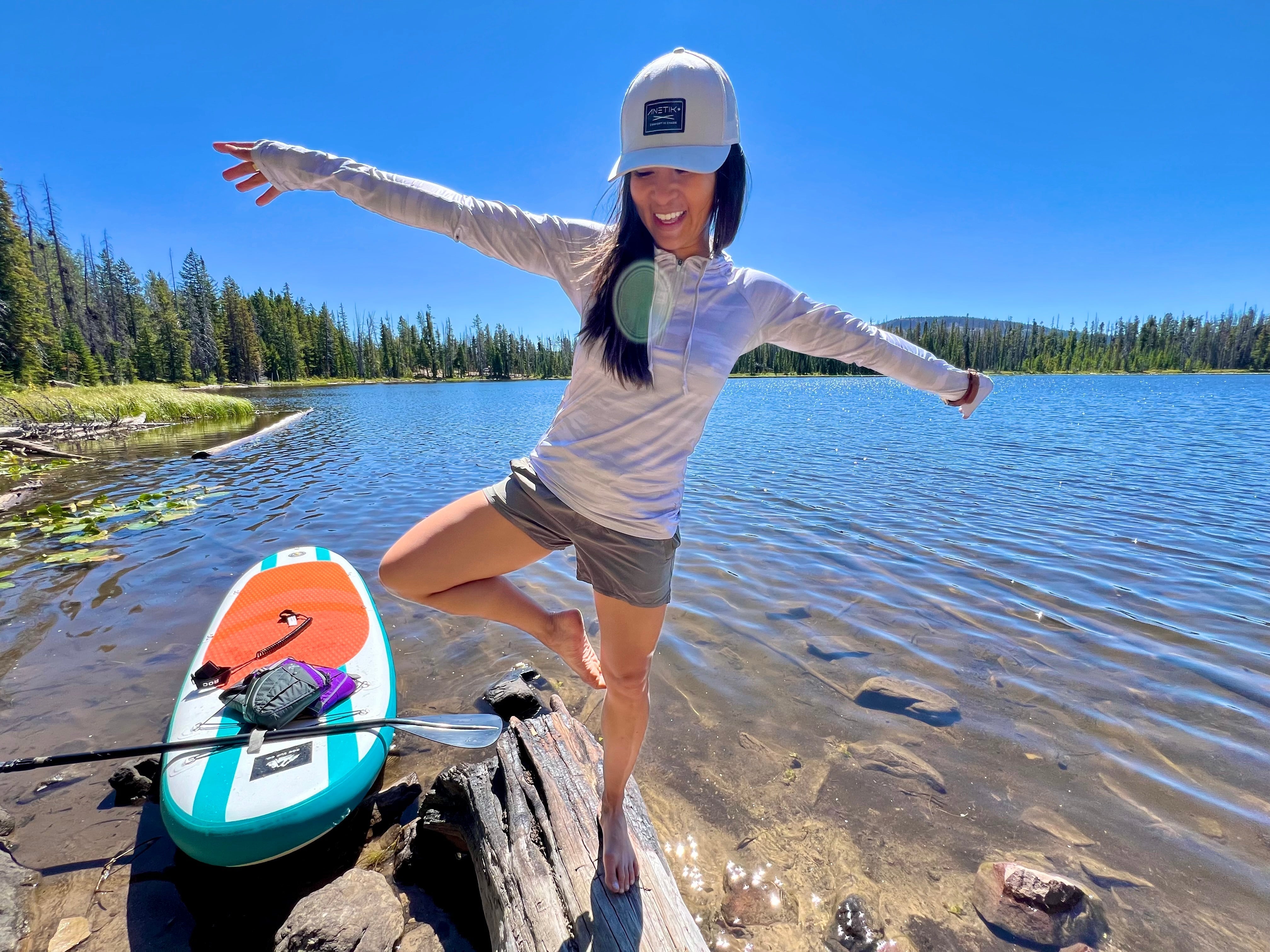 woman balancing on log with paddle board and body of water behind her