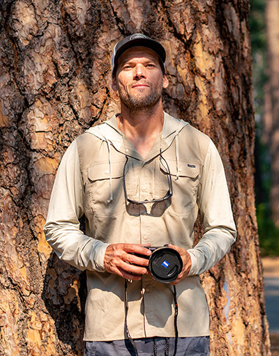 Fred Pompermayer of Anetik wearing performance outdoor shirt holding a camera while standing against a tree in a forest