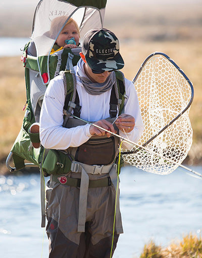 Man wearing Anetik lightweight sun protection shirt fly fishing with a child in a backpack carrier by a river