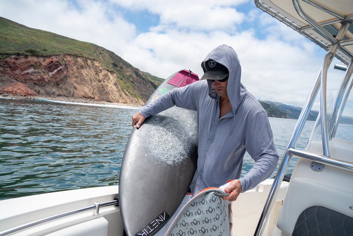 A man in a hooded sun shirt unloads surfboards from a boat near a rocky coastline.