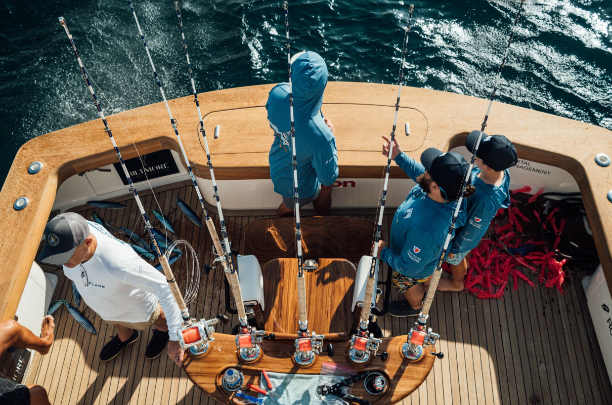 Overhead view of anglers on a sportfishing boat with rods lined up, gear on deck, and blue ocean water in the background.