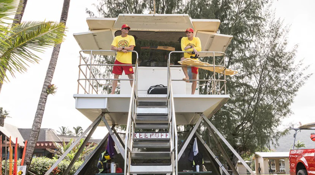 Hawaiian Lifeguard Association partners standing on a beach lifeguard tower beside palm trees and rescue gear.