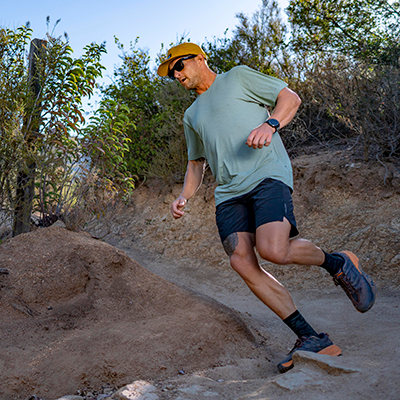 Man wearing an ANETIK performance shirt trail running down a rocky dirt path outdoors.